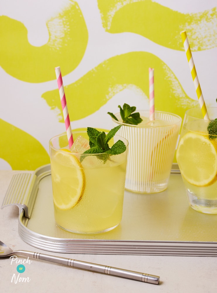 Three glasses are filled with Pinch of Nom's Homemade Lemonade, set on a silver metal tray. Each glass is decorated with a slice of fresh lemon, a colourful striped straw and a sprig of fresh mint.
