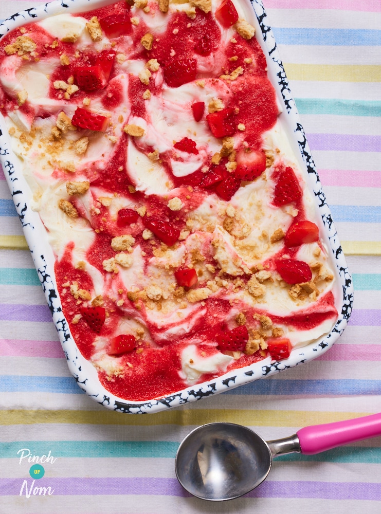 Pinch of Nom's Strawberry Cheesecake Frozen Yoghurt is served on a colourful striped tabletop in a blue and white serving dish. It's topped with strawberry puree and crushed biscuits. An ice cream scoop waits to the side to dig in.