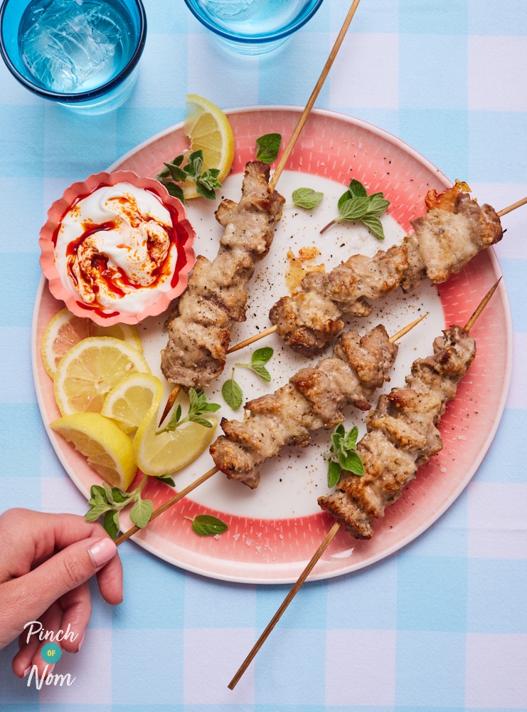 A hand is grabbing a Parmesan Pork Skewer from a red and white plate. The bamboo skewers of pork are coated in golden, melted Parmesan, served on a plate with slices of lemon, scattered herbs and a small bowl of dipping sauce. 