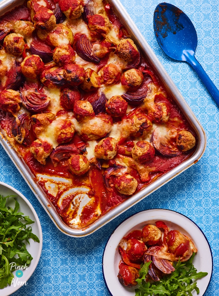 Fresh from the oven, Pinch of Nom's slimming-friendly Margherita Meatball Traybake is at the centre of a table being served onto side plates with a large spoon. The portions are plated up with a fresh, leafy green side salad.