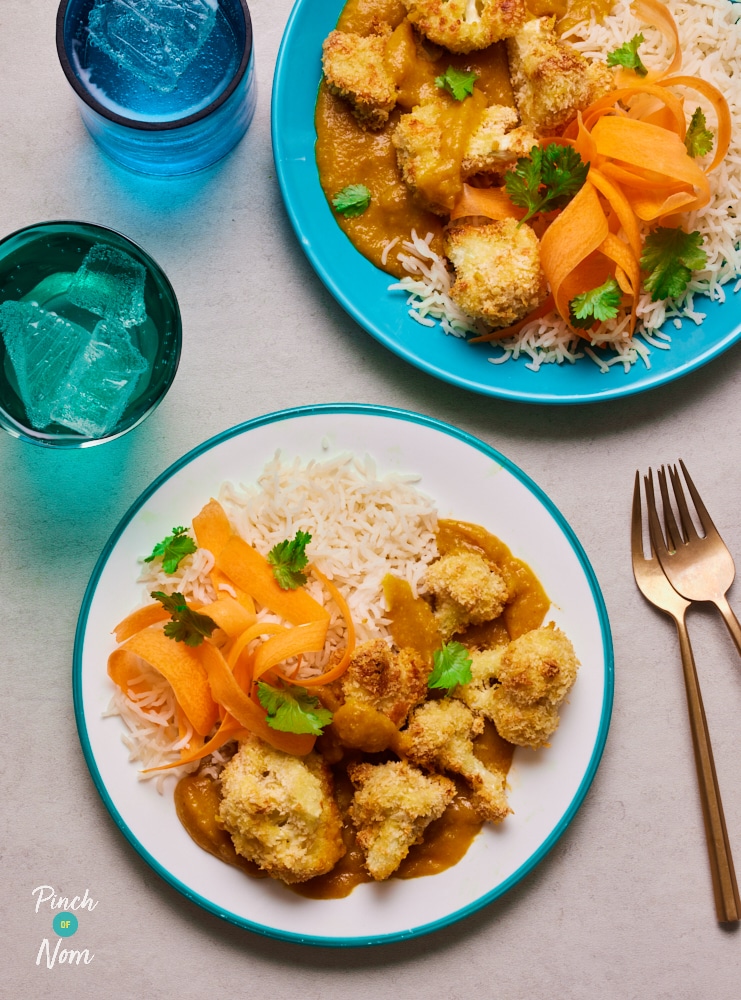 Two plates of Pinch of Nom's Caulifower Katsu sit on a kitchen counter, next to forks and glasses of ice water. The katsu curry has been served with a portion of fluffy rice and ribbons of crunchy carrot.