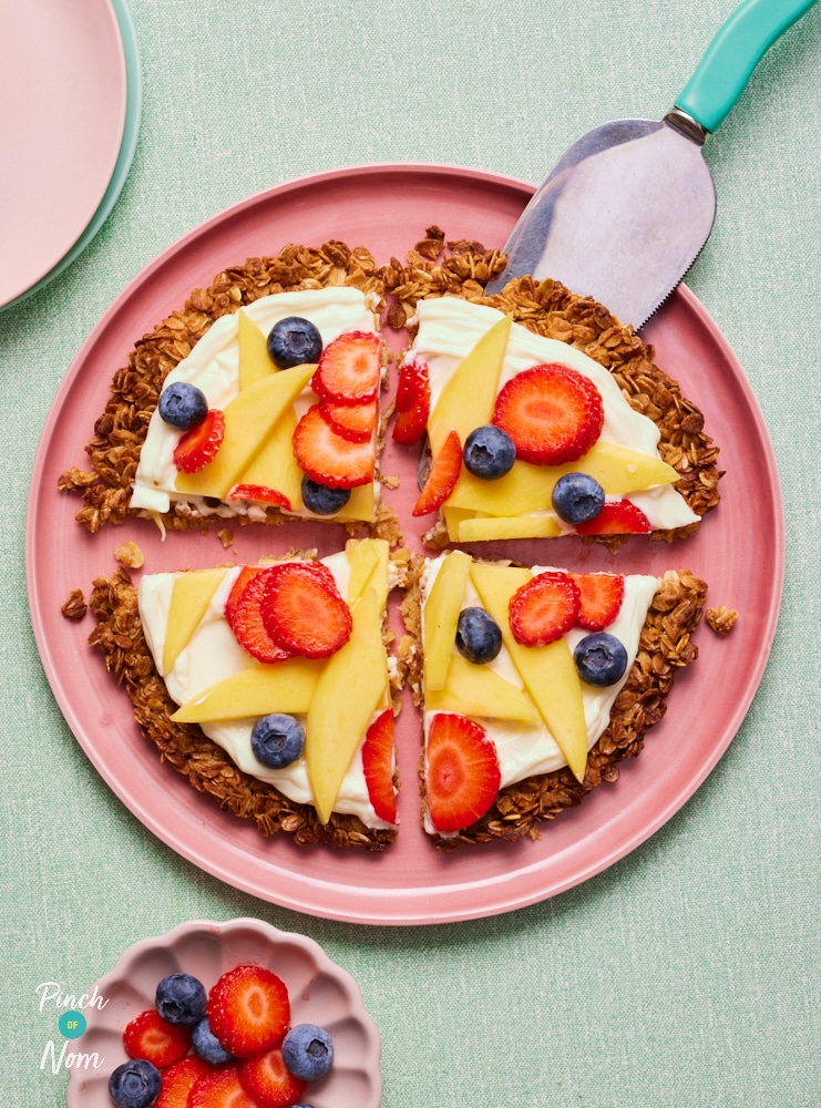 Pinch of Nom's Fruity Flapjack Pizza is plated up with a slicer reaching for one of four slices. The top is drizzled with sweet reduced-fat cream cheese, berries and mango slices.