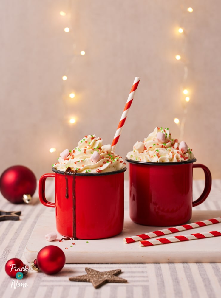 Two of Pinch of Nom's Christmas Hot Chocolates are served on a festive-looking table in cosy red mugs, with baubles and fairy lights scattered around. Both hot chocolates are topped with cream, marshmallows and sprinkles.