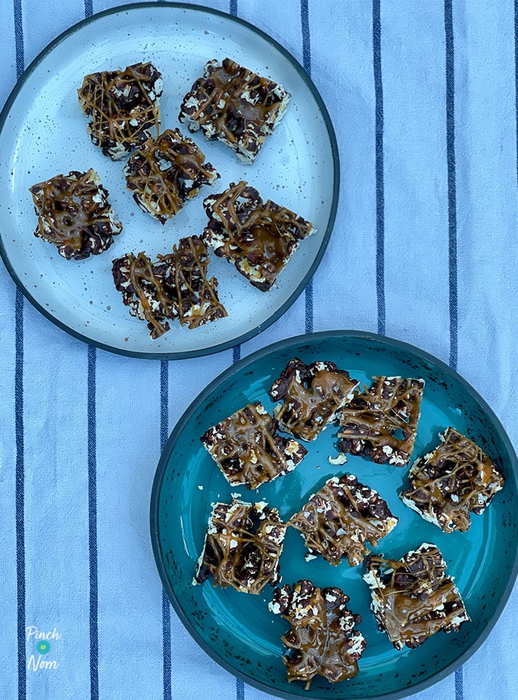 A table is set with a white striped tablecloth and two blue plates, filled with Pinch of Nom's Salted Caramel and Chocolate Popcorn Bites. The chocolate and popcorn mixture has been cut into squares, and each one os drizzled with salted caramel.