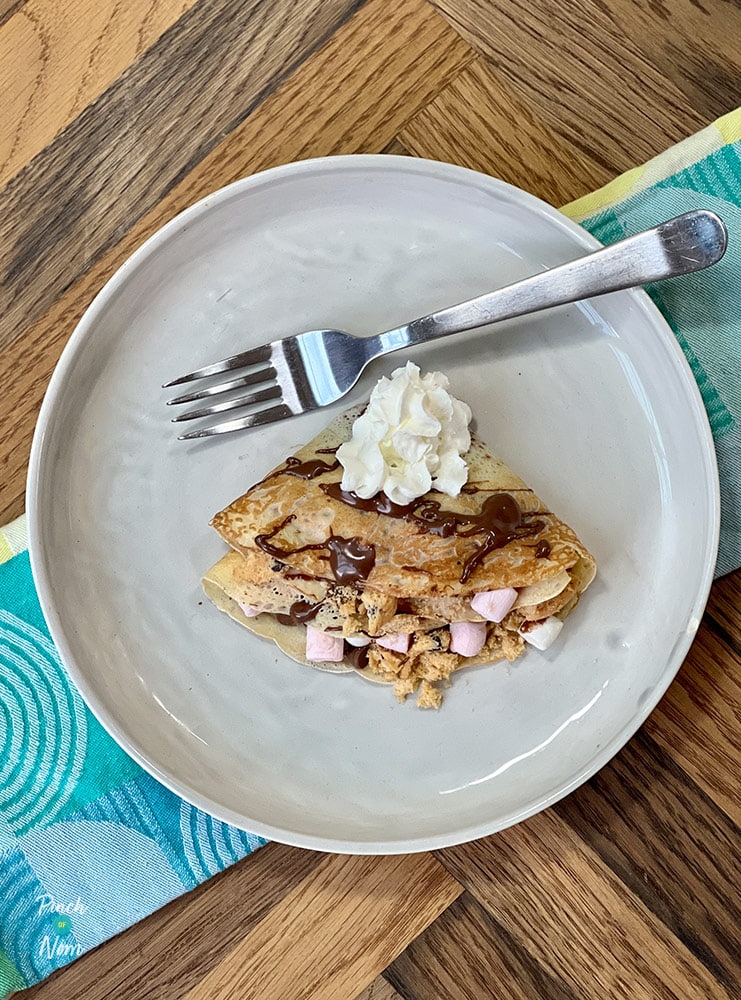 A table is set with a plate of Pinch of Nom's slimming-friendly S'mores Pancakes; the pancake is folded over and topped with whipped cream and a chocolate drizzle, while a fork waits to tuck in.