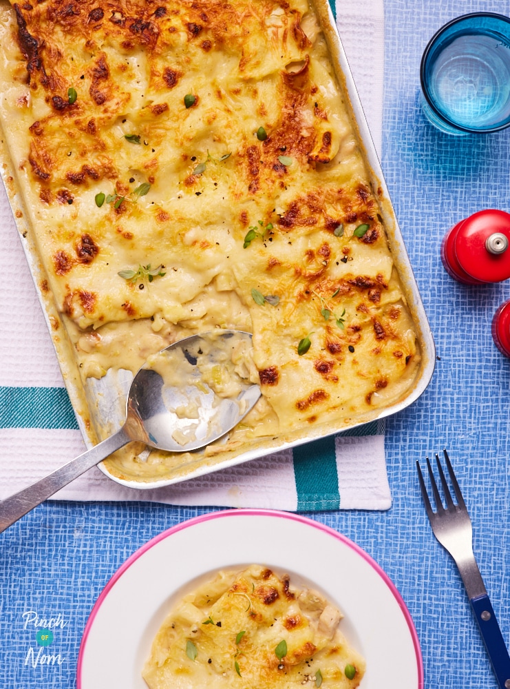 Pinch of Nom's Chicken, Mushroom and Leek Lasagne is served freshly baked from the oven with a gooey golden topping of cheese in an oven dish. A portion has been scooped from the dish and is served up on a white plate to the side, with a fork waiting to tuck in.