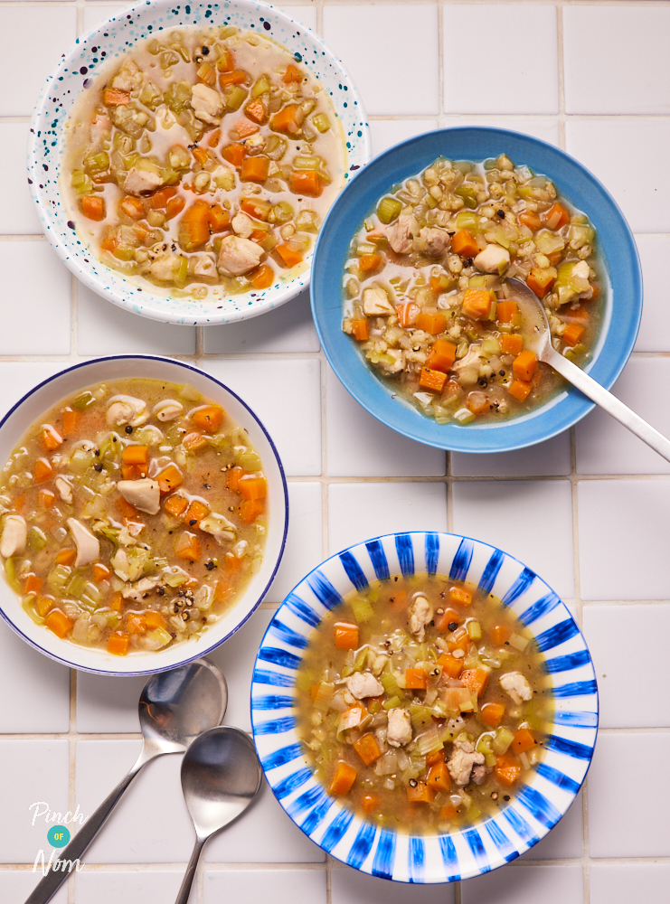 Four portions of Pinch of Nom's Chicken, Leek and Barley Soup are plated up in colourful bowls on a white tiled kitchen counter. There are spoons waiting to tuck in.