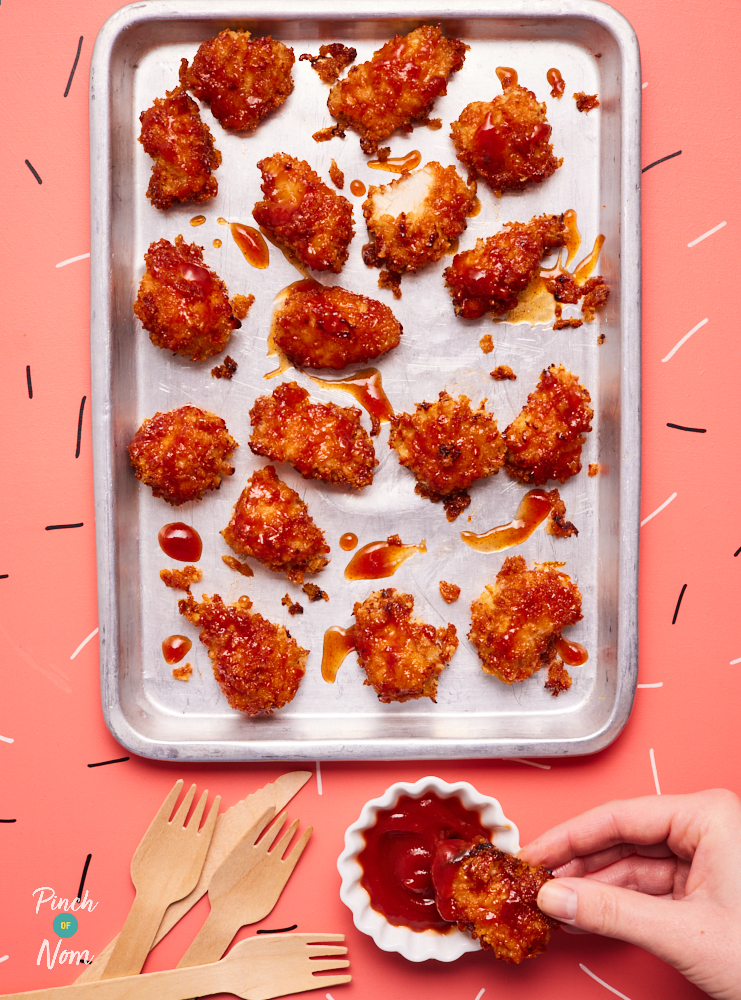 A baking tray is filled with Pinch of Nom's Sticky Chicken Nuggets. The crispy nuggets are coated in a sweet, sticky sauce and served with a dipping pot of ketchup. Wooden forks are laid out on the table and a hand holds one nugget, dipping it into the sauce.