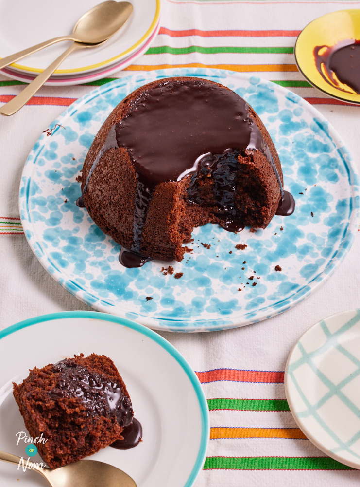 A table is set with a white and multi-coloured striped tablecloth. In the centre, a light blue patterned plate is filled with a dome-shaped chocolate cake, with chocolate sauce poured over the top and running down the sides. A slice has been cut from the front of the cake and served on a small white plate that can be seen in the bottom left of the photo. This is Pinch of Nom's slimming-friendly Chocolate Sponge Pudding.