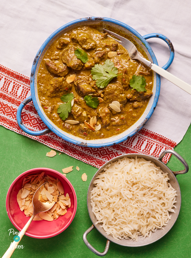 A green table is laid with a white cloth, with a red embroidered border. A bowl of Pinch of Nom's Lamb Pasanda is on the table, ready to eat, garnished with coriander. A fork is resting in the dish. A small bowl of fluffy rice is nearby, with a smaller bowl of toasted almonds ready to sprinkle on top.