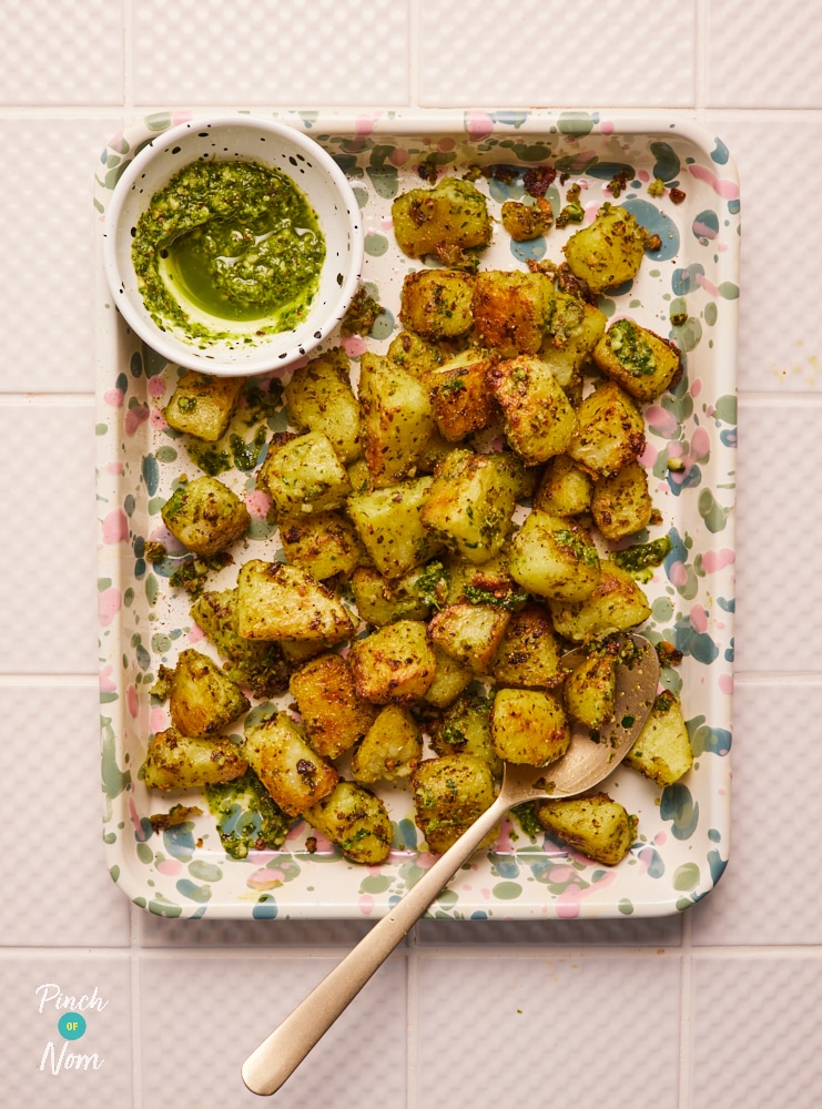 A colourful ceramic oven tray is filled with Pinch of Nom's Pesto Roast Potatoes. The golden roasted potatoes are covered in herby green pesto, with a pot of extra pesto nearby, and a spoon ready to serve.