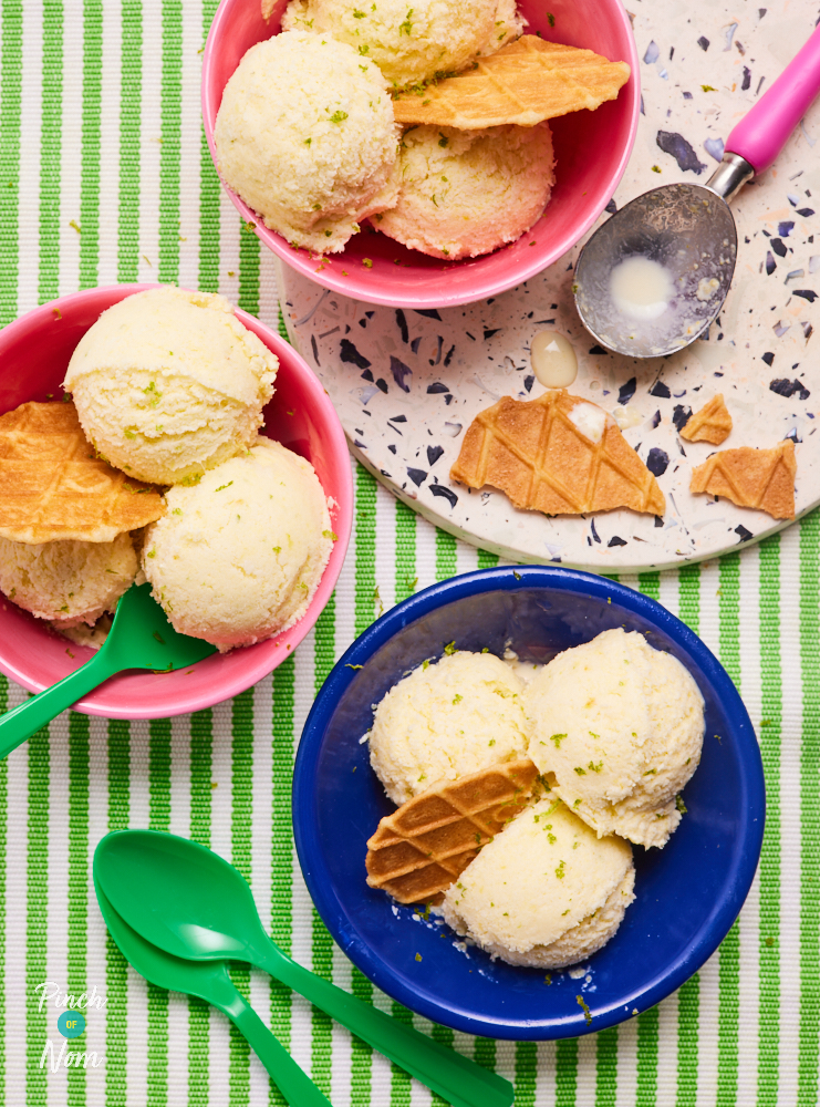 A table is laid with a green and white tablecloth and 3 bowls of Pinch of Nom's Piña Colada Ice Cream. Each colourful bowl has three scoops of ice cream and a thin, round wafer. Bright green spoons are nearby, ready to dig in.
