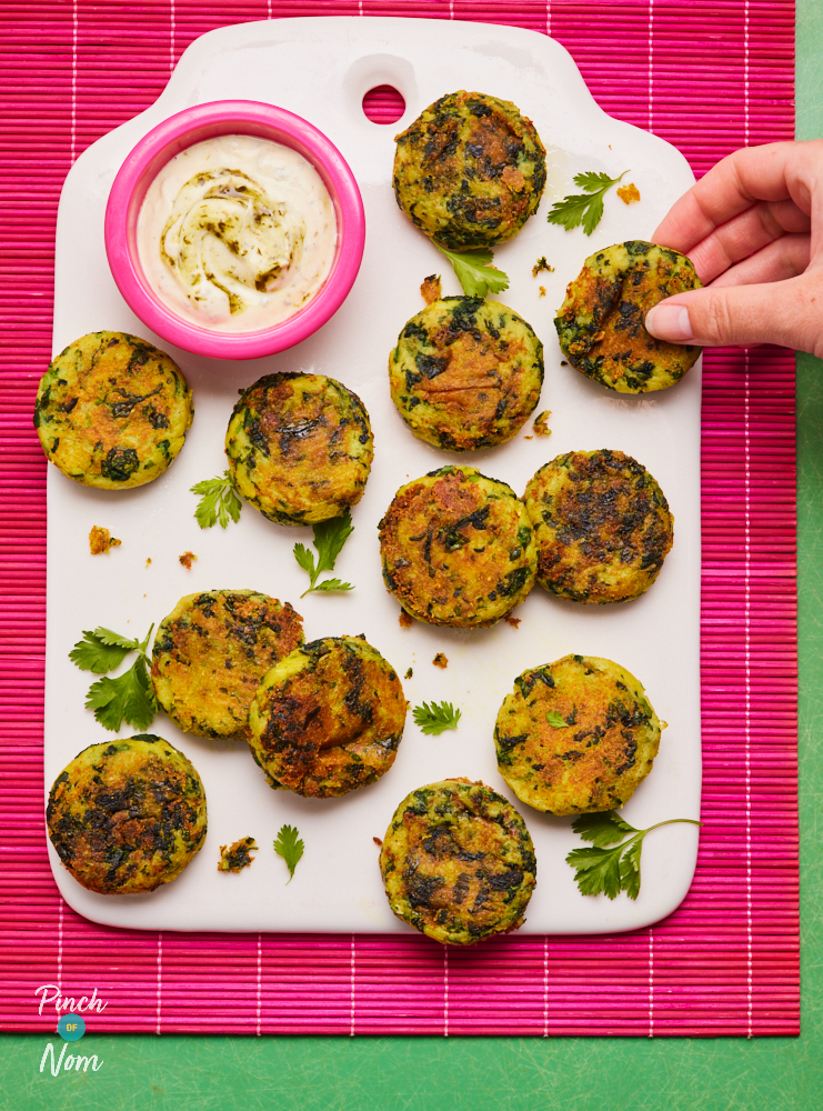 A serving board is filled with a batch of crispy-looking Saag Aloo Fritters waiting to be dipped into a homemade mint-infused yoghurt to the side. A hand is reaching to grab one of the fritters!