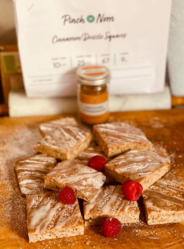 A chopping board is topped with a pile of Pinch of Nom's slimming-friendly Cinnamon Drizzle Squares. They're topped with cinnamon drizzle, icing dust and fresh raspberries.