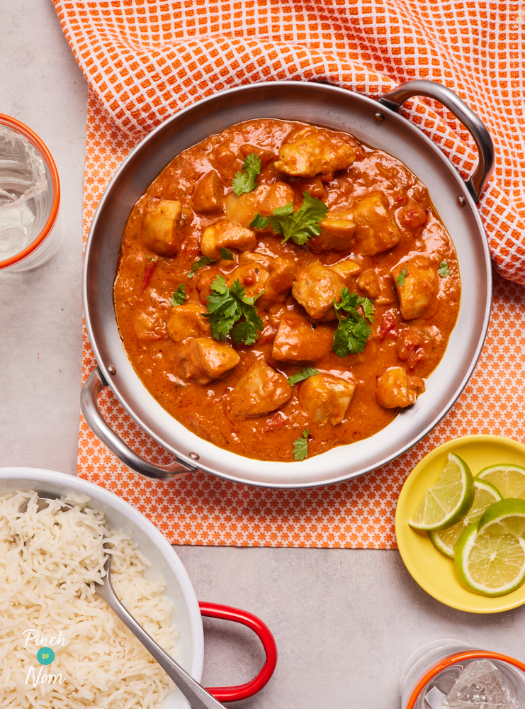 Pinch of Nom's Spicy Coconut and Lime Chicken Curry served in a large pan, covered with a garnish of coriander. Lime wedges are ready to squeeze on a yellow side plate, and basmati rice is ready to be served from a white bowl.
