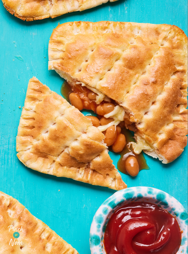 A close-up image of one of Pinch of Nom's Cheesy Baked Bean Bakes where the cheesy bean filling is spilling out from the middle – a small pot of ketchup waits to the side to be dipped.