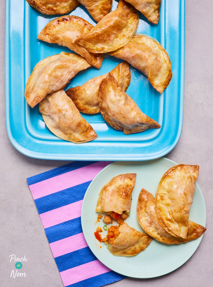 A baking tray of Empanadas is fresh out of the oven, and three Empanadas have been served onto a small pale green plate. The Empanadas are golden brown and one has been cut in half to reveal the vegetable filling.