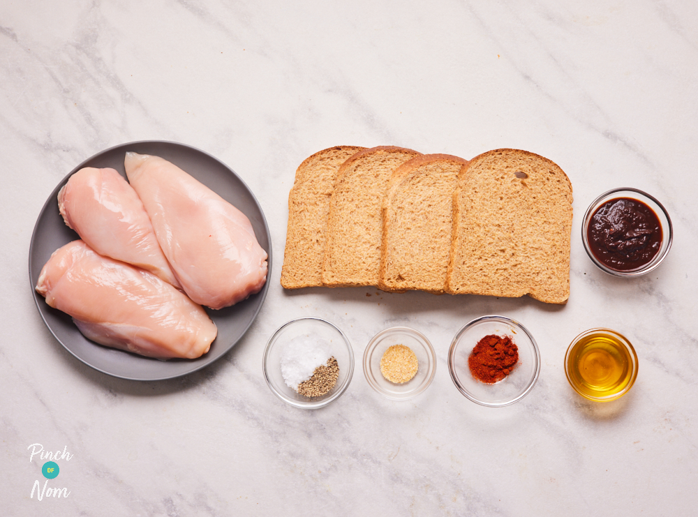 The ingredients for Pinch of Nom's Chipotle Popcorn Chicken are laid out on a kitchen counter. There are 3 chicken breasts, slices of wholemeal bread, honey, chipotle paste and other herbs and seasonings.