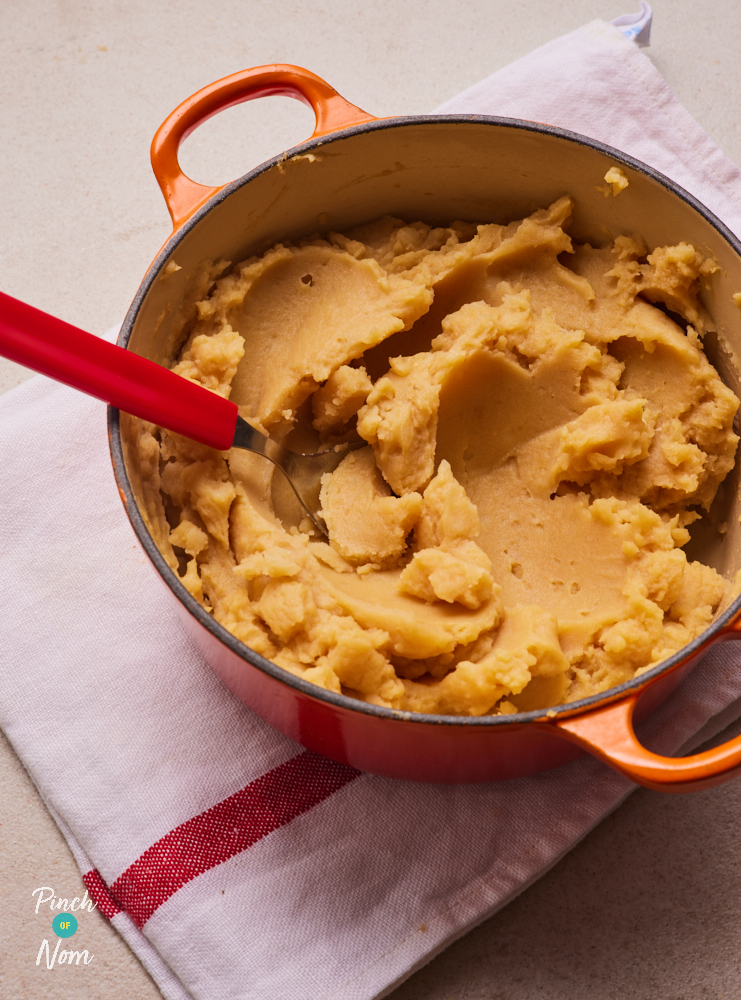 Pinch of Nom's Marmite Mash is served in a large, red, ovenproof dish with a red serving spoon waiting to scoop a serving out.