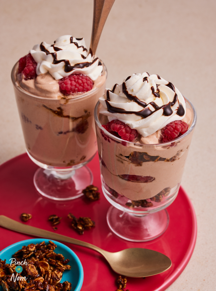 Two glasses of Pinch of Nom's Raspberry Mocha Granola pots are served, topped with a swirl of cream, raspberries and a drizzle Sweet Freedom Choc Shot. Extra granola waits to the side to be sprinkled on top.