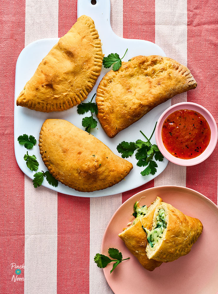 A plate serves three of Pinch of Nom's Air Fryer Cheesy Garlic Naanzone with a small pot of chutney to the side for dipping. A half-eaten naanzone rests on a side plate with the gooey middle spilling out.