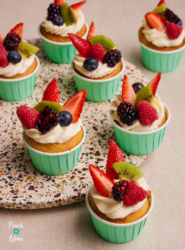 Pinch of Nom's Fruity Cupcakes are served in colourful cases on a speckled serving plate topped with strawberry, kiwi, blueberry and raspberry slices.