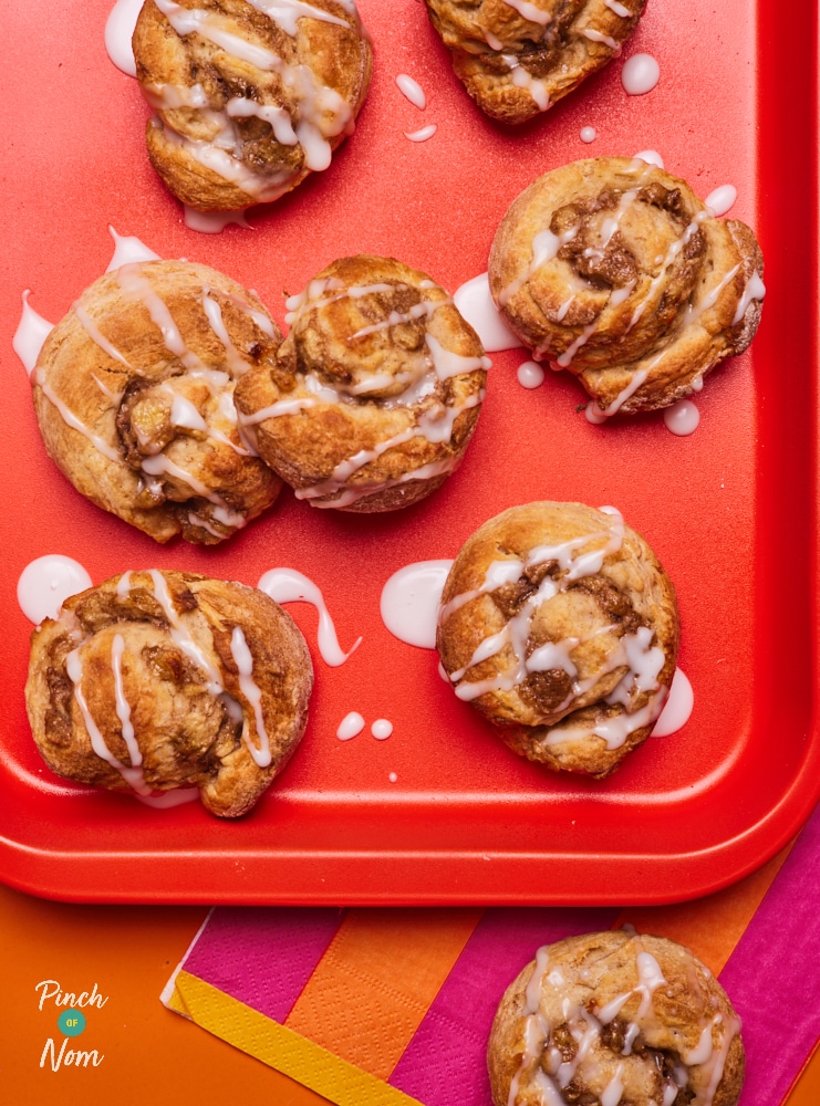 On a bright-red tray, a freshly-baked batch of Pinch of Nom's Banana Bread Cinnamon Rolls are laid out, drizzled with a generous amount of icing sugar. The sweet banana and cinnamon filling is bursting from the centre of each roll.