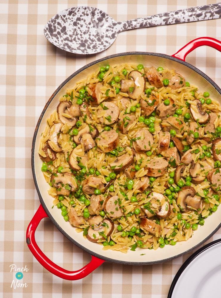 A large ovenproof casserole dish is filled with Pinch of Nom's slimming-friendly Herby Sausage Orzo, served at the centre of a set table ready to begin dishing out.