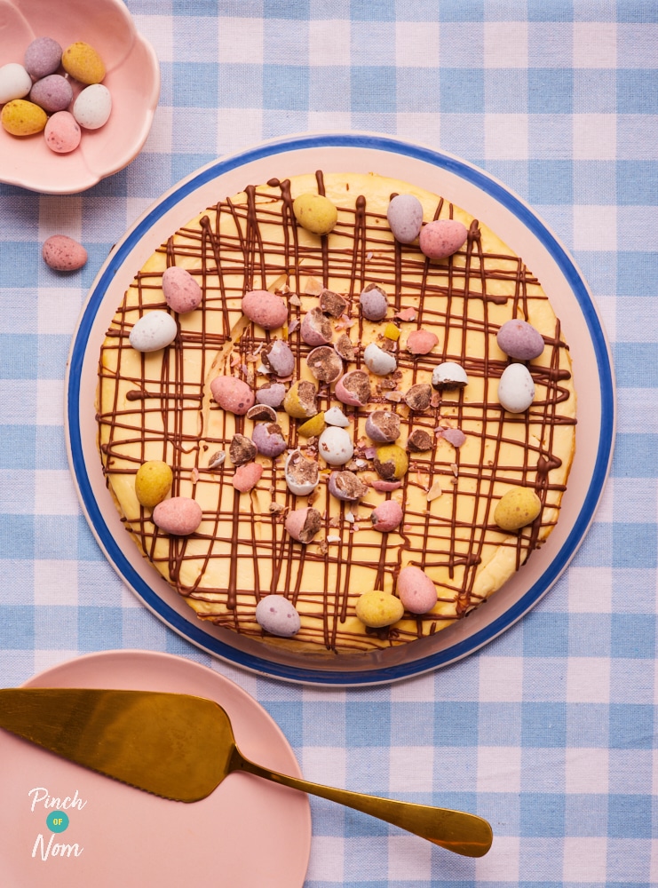 At the centre of a gingham table, Pinch of Nom's slimming-friendly Mini Egg Cheesecake is served decorated with a chocolate drizzle and a garnish of mini eggs, ready to be sliced.