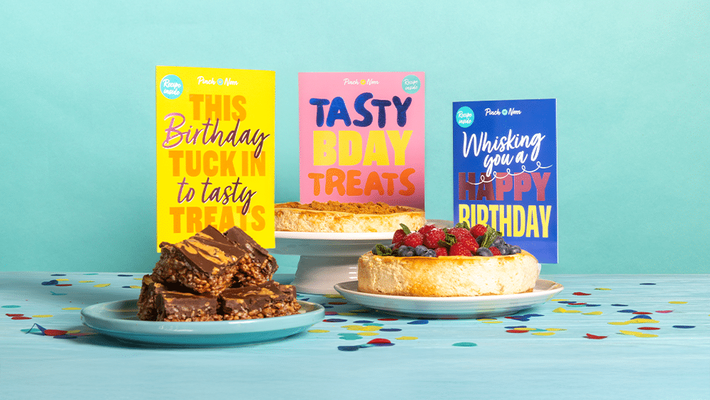Pinch of Nom's Birthday Cards are stood side by side on a party table spread featuring the three recipes that come with each card: Chocolate Peanut Butter Bars, Biscoff Cheesecake and Raspberry and Blueberry Baked Cheesecake.
