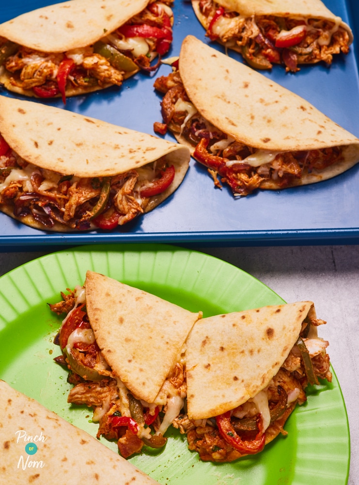 Four of Pinch of Nom's slimming-friendly Chicken Tacos are served filled with shredded chicken, peppers, and melted cheese on a blue baking tray, with three more tacos served on a bright green plate below.