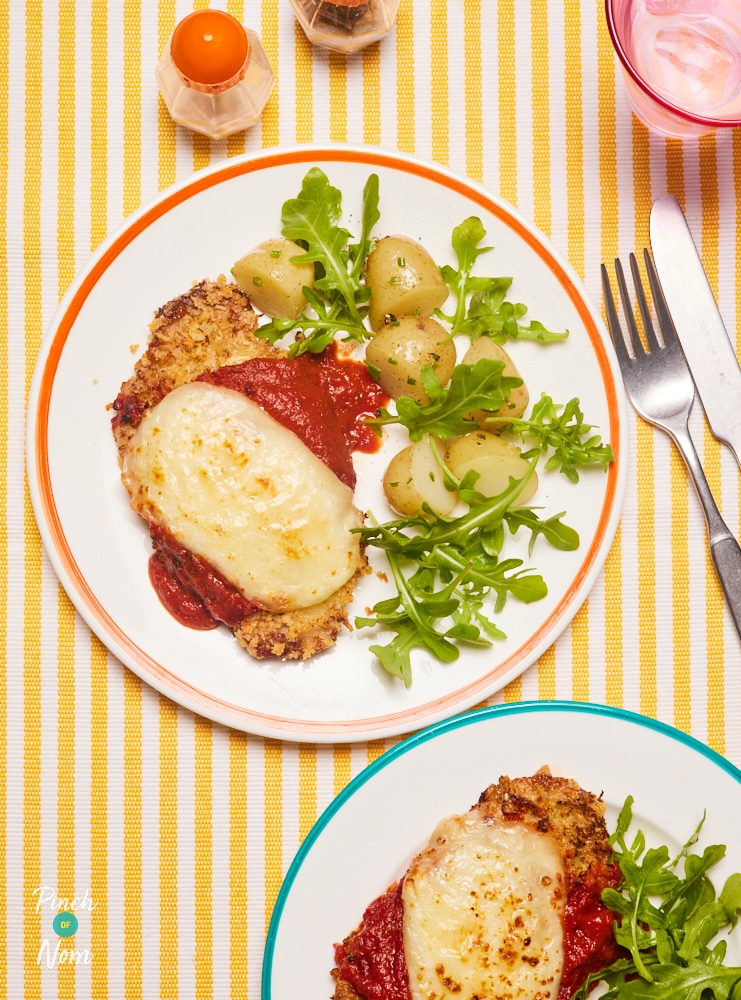 A table is set with two plates of Pinch of Nom's slimming-friendly Air Fryer Pork Parmigiana recipe. The cheesy-topped escalopes are served beside a green salad and new potatoes, with a set of cutlery to one side ready to dig in.