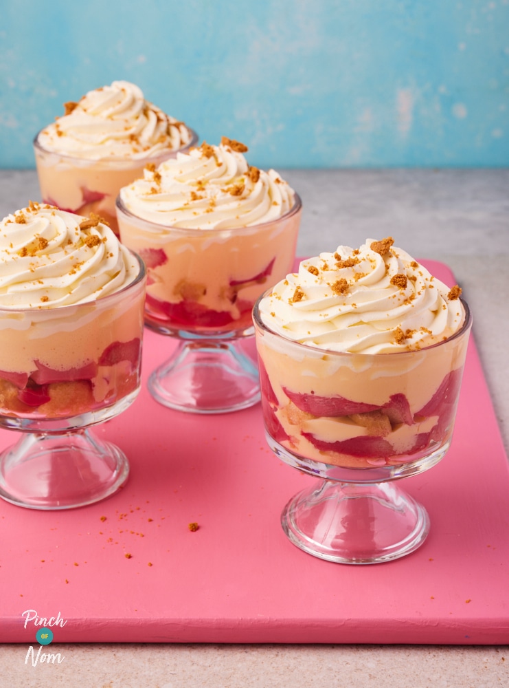 A table is set with four dessert dishes, filled with Pinch of Nom's slimming-friendly Rhubarb and Custard Trifles. The trifles are served in clear dessert glasses, revealing the bright-colours layers of yellow custard and pink rhubarb.