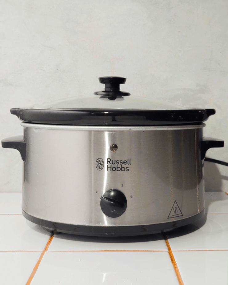 A sparkling clean Russell Hobbs slow cooker with a glass lid sits on a white tiled surface.