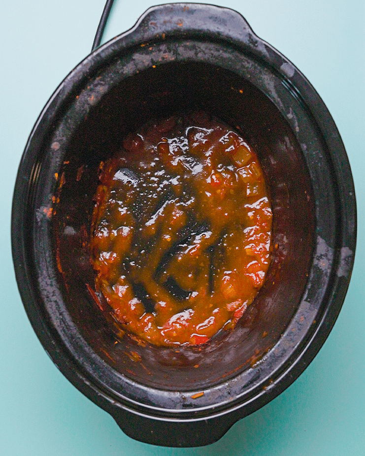 A top-down view into the black ceramic pot of a slow cooker. The pot contains remnants of a cooked dish, appearing to be a tomato-based sauce with some solid pieces. The sauce is coating the bottom and sides of the pot.