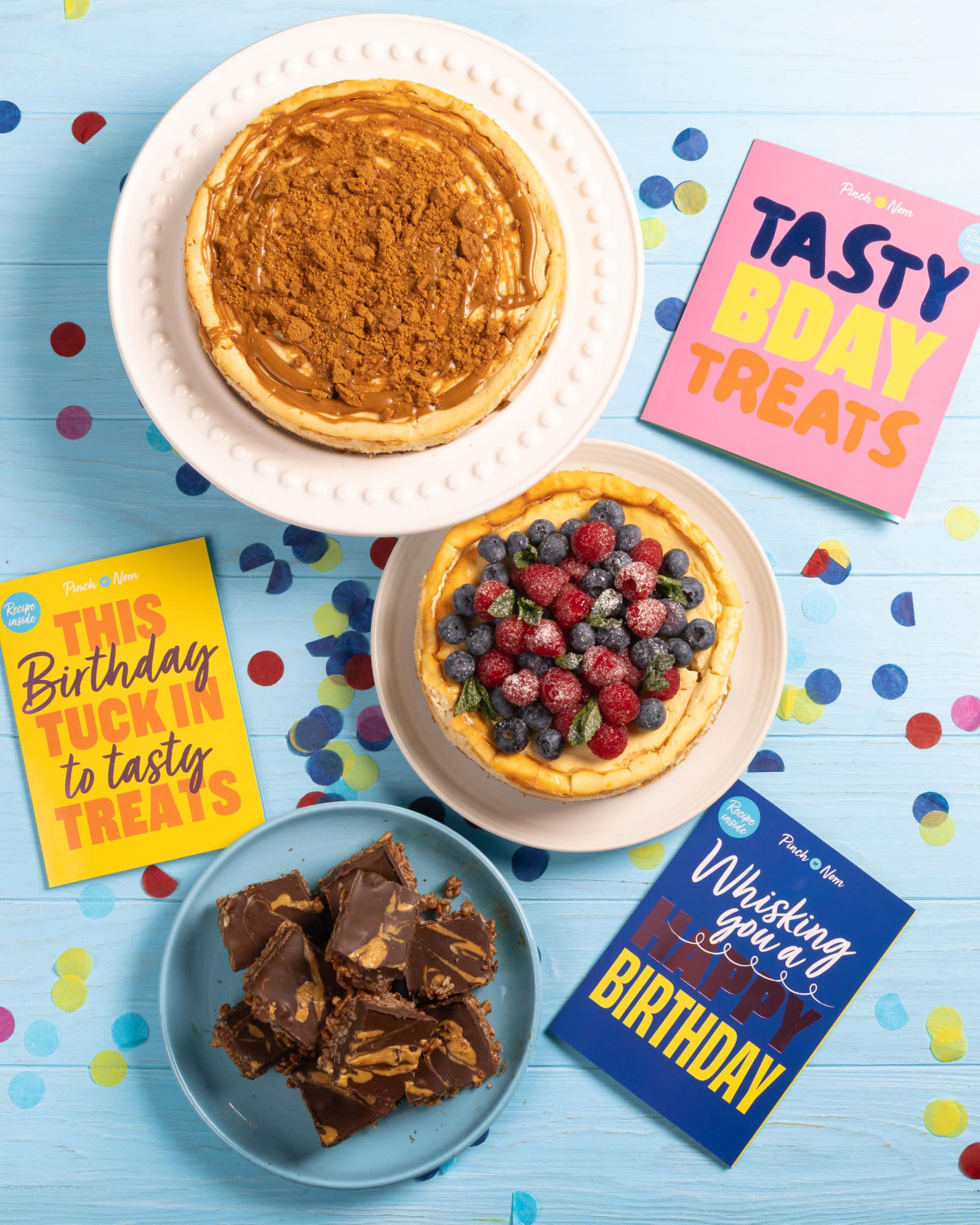 A table is laid with Pinch of Nom's Birthday Cards and confetti, alongside the recipes featured in each card: Chocolate Peanut Butter Bars, Biscoff Cheesecake and Raspberry and Blueberry Baked Cheesecake.