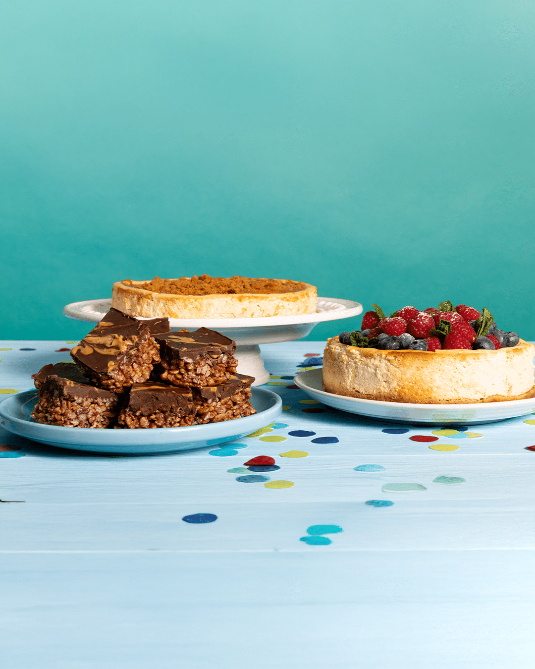 A party table is set with the recipes from Pinch of Nom's Birthday Cards: Chocolate Peanut Butter Bars, Biscoff Cheesecake and Raspberry and Blueberry Baked Cheesecake.