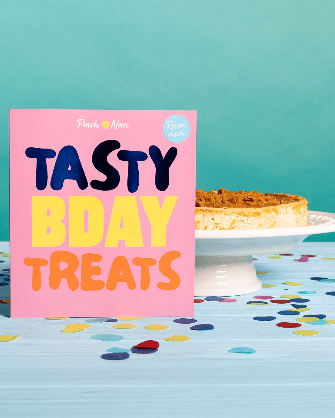 Pinch of Nom's 'Tasty Bday Treats' card is stood on a party table decorated with confetti alongside Pinch of Nom's Biscoff Cheesecake recipe.