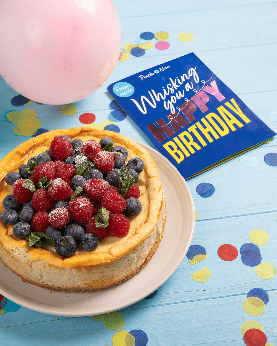 Pinch of Nom's 'Whisking You a Happy Birthday' card is laid on a party table decorated with confetti and balloons alongside Pinch of Nom's Raspberry and Blueberry Baked Cheesecake recipe.