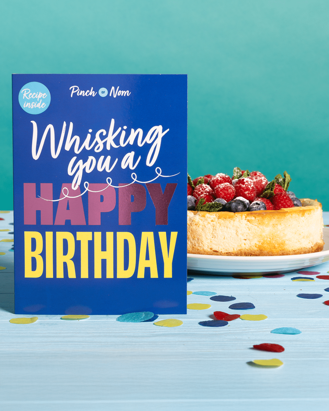 Pinch of Nom's 'Whisking You a Happy Birthday' card is stood on a party table decorated with confetti alongside Pinch of Nom's Raspberry and Blueberry Baked Cheesecake recipe.