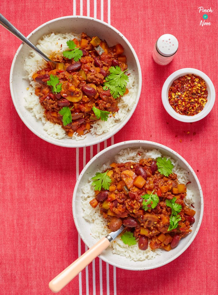 Two bowls of Slow Cooker Pinch of Noms Chilli Con Carne are served on a red tabletop. Both portions are topped with fresh coriander leaves; a small pot of chilli flakes waits to one side, waiting to be scattered over.