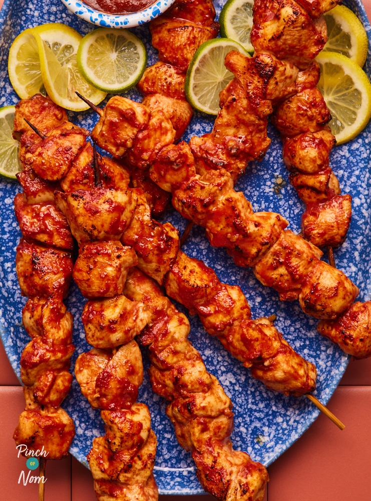 A large serving plate is filled with Pinch of Nom's slimming-friendly Lemonade Chicken Skewers, with lemon wedges for squeezing on top and a small pot of banana ketchup for dipping.