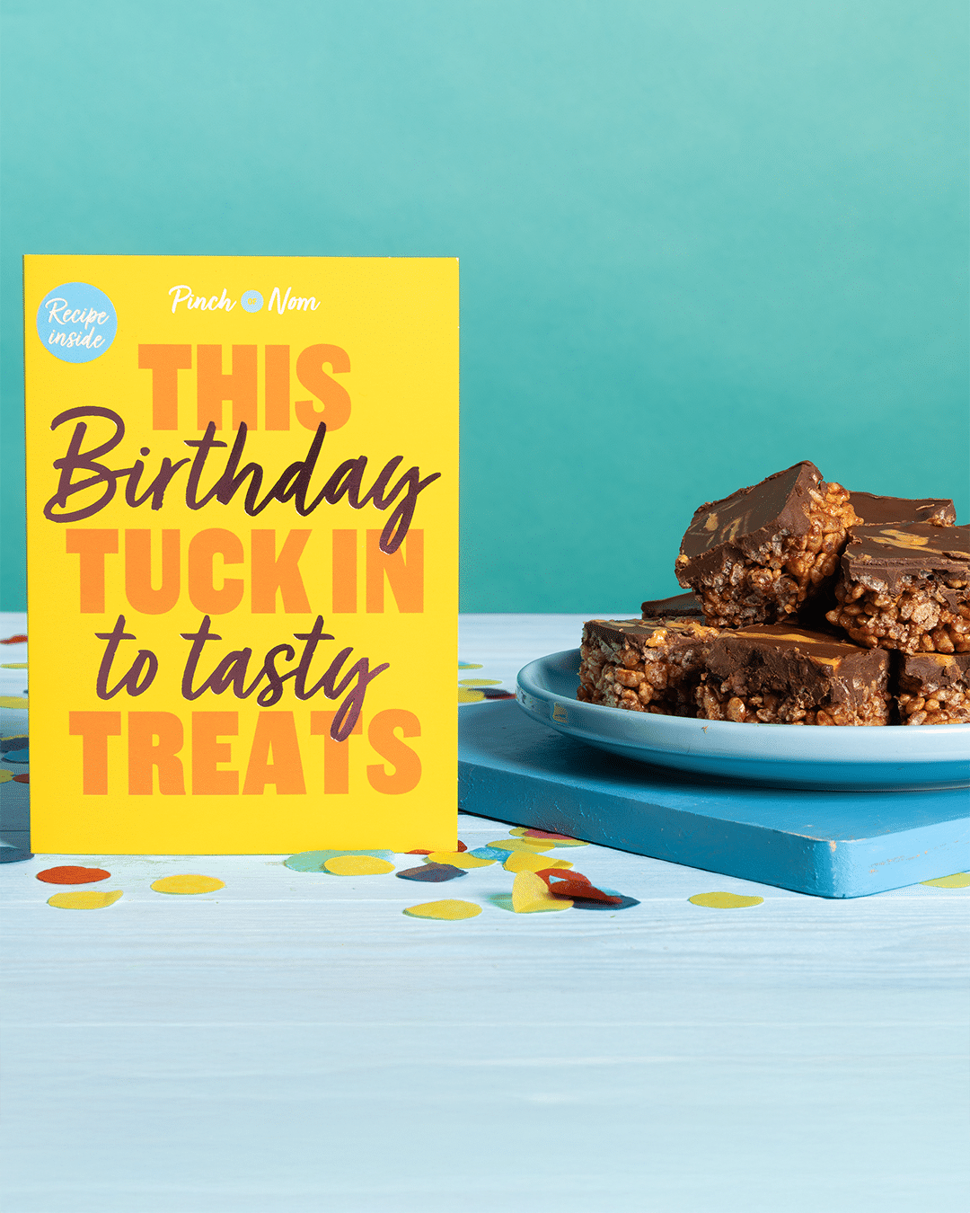 Pinch of Nom's 'This Birthday Tuck Into Tasty Treats' card is stood on a party table decorated with confetti alongside a plate of Pinch of Nom's Chocolate Peanut Butter Bars.