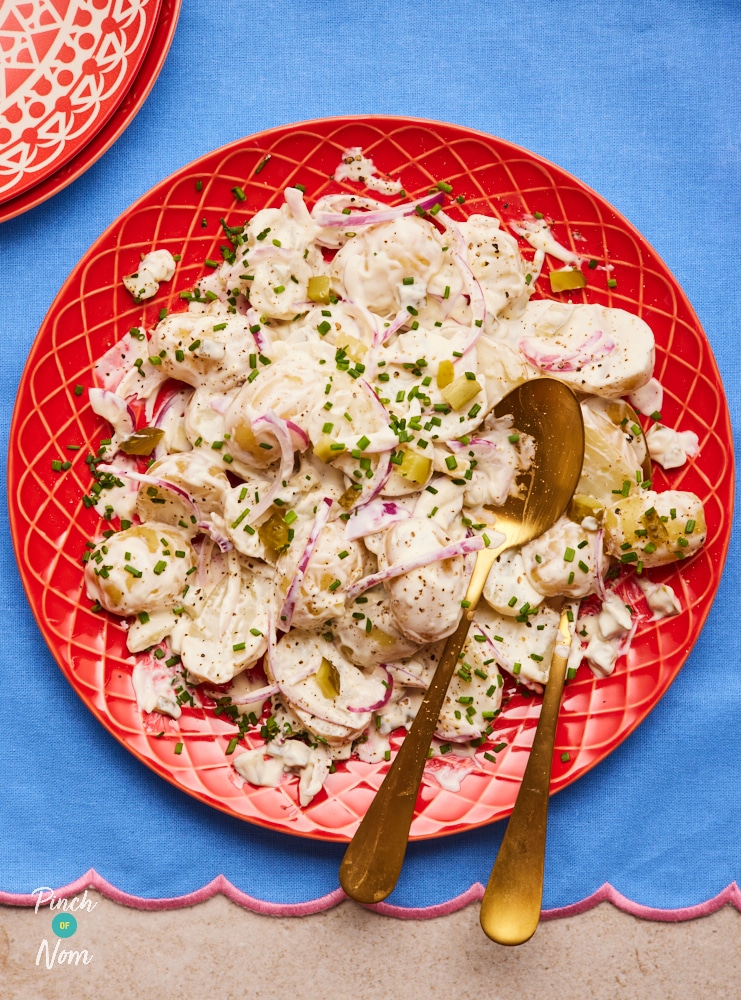 A table is set with Pinch of Nom's slimming-friendly Pickle Potato Salad served on a bright red plate, seasoned with salt and pepper, with finely sliced chives sprinkled on top.