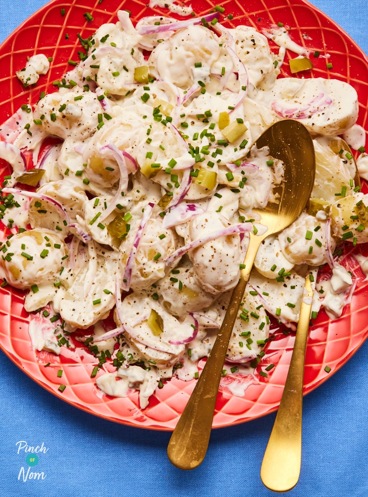 A table is set with Pinch of Nom's slimming-friendly Pickle Potato Salad served on a bright red plate, seasoned with salt and pepper, with finely sliced chives sprinkled on top.