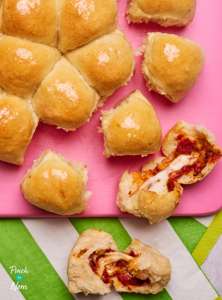A bright pink serving board is topped with a freshly-baked batch of Pinch of Nom's slimming-friendly Pizza Dough Balls. One of the dough balls has been ripped apart, revealing the gooey tomato and cheese centre.