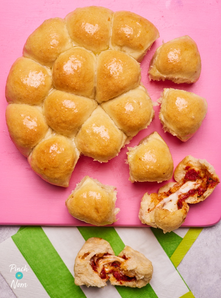 A bright pink serving board is topped with a freshly-baked batch of Pinch of Nom's slimming-friendly Pizza Dough Balls. One of the dough balls has been ripped apart, revealing the gooey tomato and cheese centre.