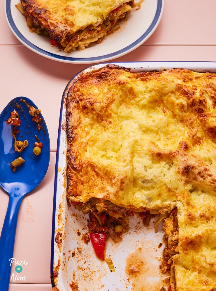 Pinch of Nom's slimming-friendly Summer Vegetable Lasagne is served freshly-baked in a large oven dish; one portion has been dishes onto a side plate by a large blue spoon, revealing the tomatoey, veg-filled centre.